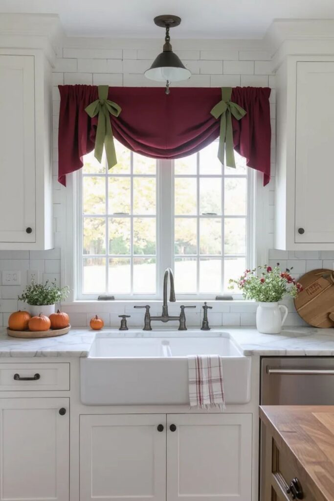 White farmhouse kitchen with burgundy fabric valance featuring sage green ribbon ties over sink window, fall styling with small pumpkins and flowers on marble countertops, subway tile backsplash, creating cozy minimalist fall kitchen decor