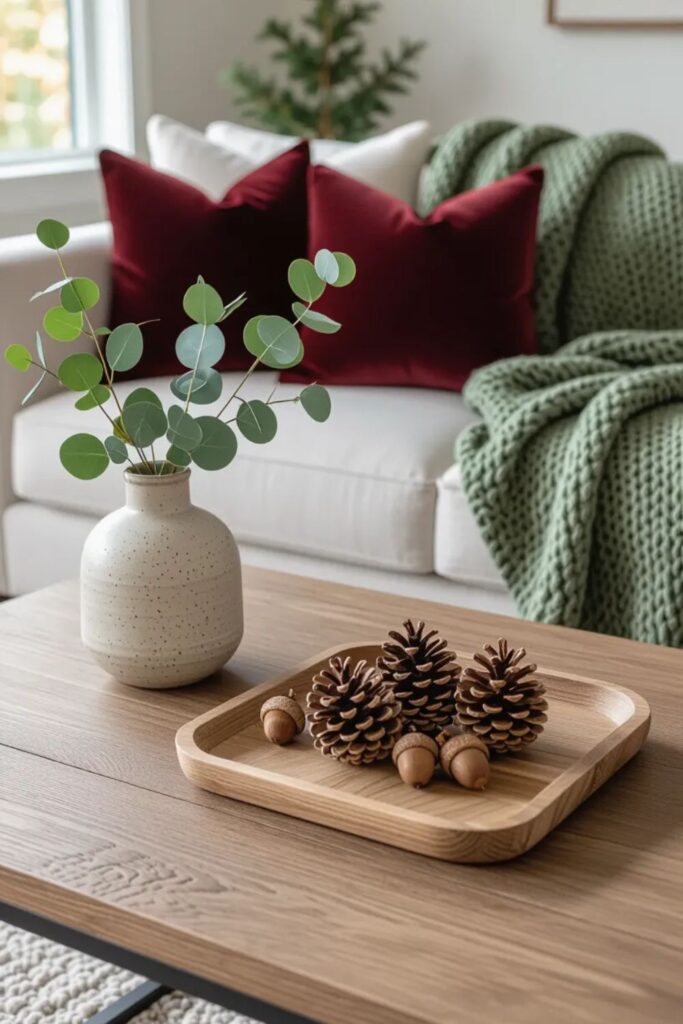 Minimalist living room with burgundy velvet throw pillows and sage chunky knit blanket on neutral sofa, wooden coffee table with natural fall elements including pinecones and acorns on wood tray, eucalyptus in ceramic vase, layered seasonal textures for cozy fall home decor
